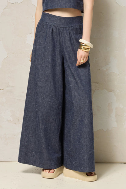 Waist down view of a woman wearing the Rama Pants by Cokluch in Indigo Speckle, textured denim pants with wide legs and a smooth elastic waistband. She is wearing them with a matching top and standing in front of a grey wall. 