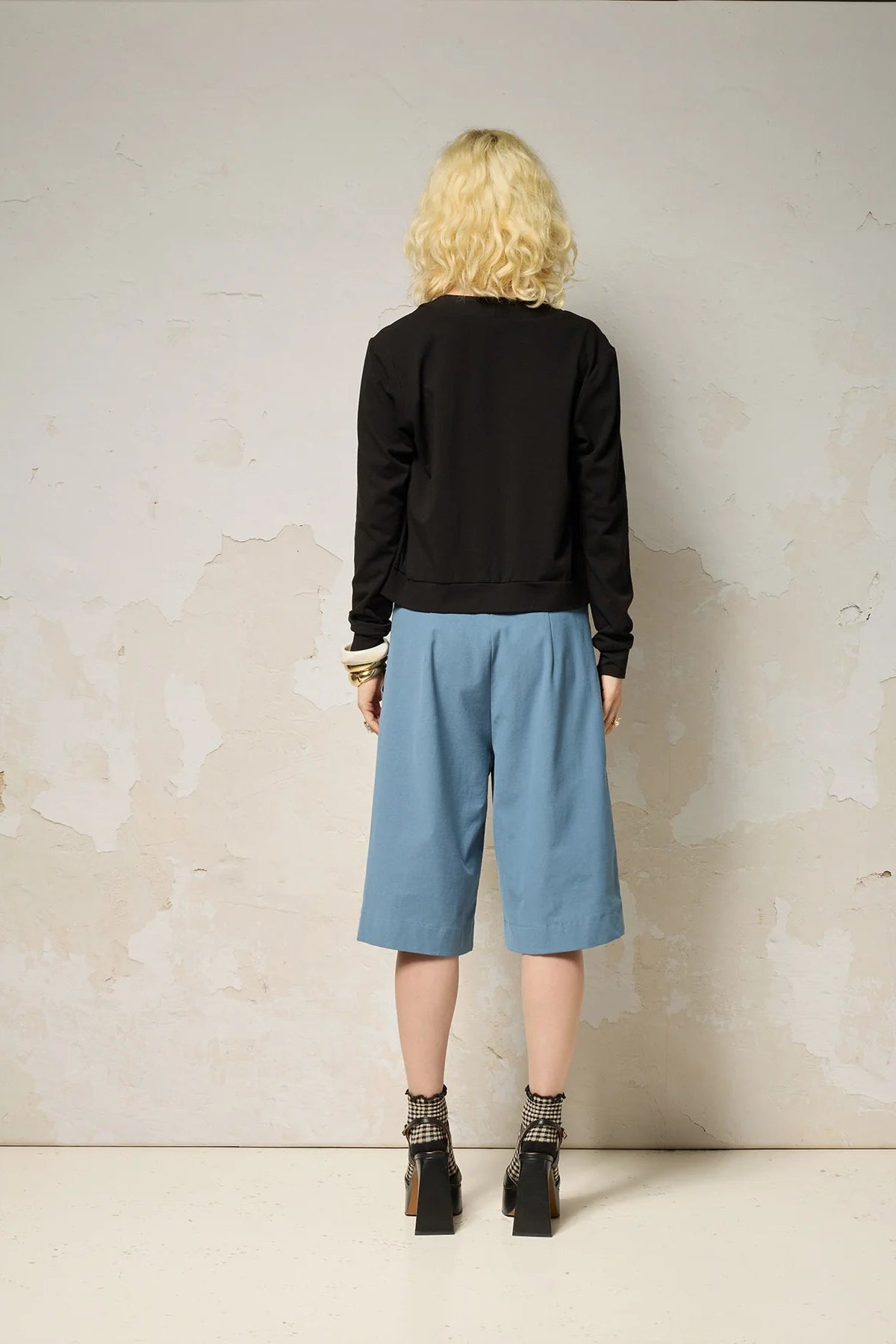 Back view of a woman wearing the Zephyr Cardigan by Cokluch in Black, a long sleeved waist length cardigan with a v-neck and four buttons. She is wearing it with the Hebe striped tank and blue shorts against a plain background. 