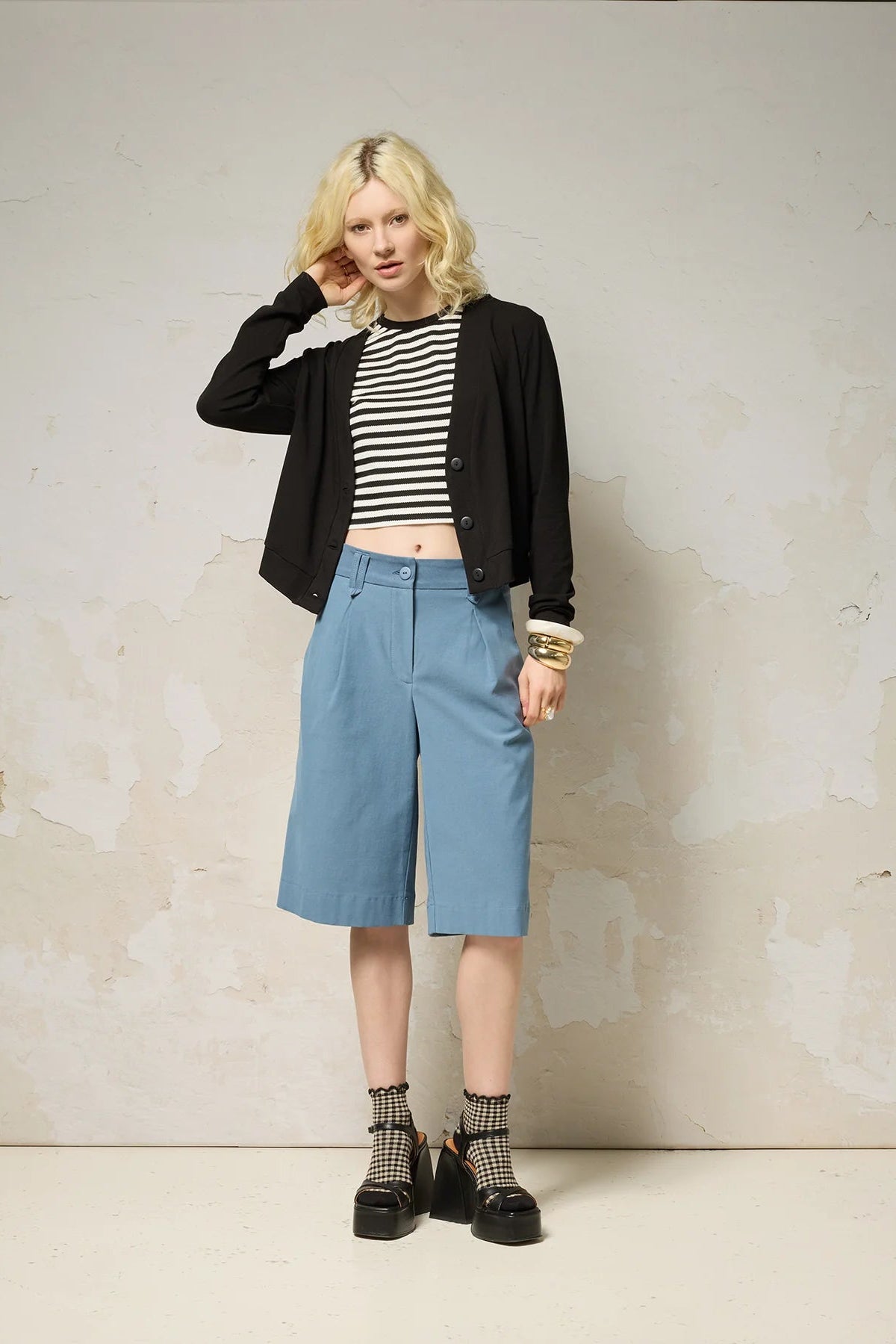 A woman wearing the Zephyr Cardigan by Cokluch in Black, a long sleeved waist length cardigan with a v-neck and four buttons. She is wearing it with the Hebe striped tank and blue shorts against a plain background. 