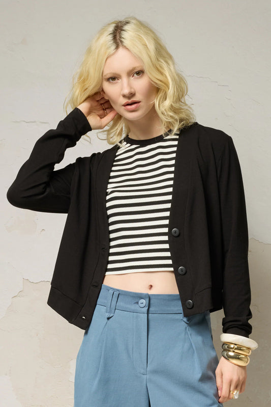 Waist up view of a woman wearing the Zephyr Cardigan by Cokluch in Black, a long sleeved waist length cardigan with a v-neck and four buttons. She is wearing it with the Hebe striped tank and blue shorts against a plain background. 