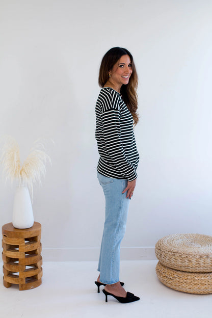 Side view of a woman wearing the Munro Bamboo Crewneck sweater by Ang Hill in black and white stripes. She is wearing it with light blue jeans against a white background.
