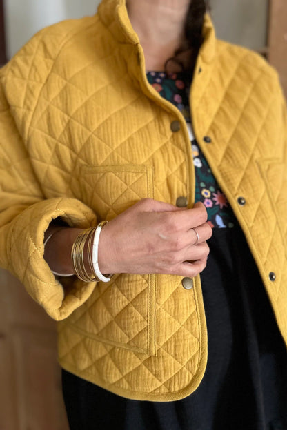 A close up of a woman wearing the Artemesia Jacket by Kazak in Mustard, a quilted jacket with snaps up the front and large patch pockets. She is wearing it over  the Lavendula Dress in Farmland. 