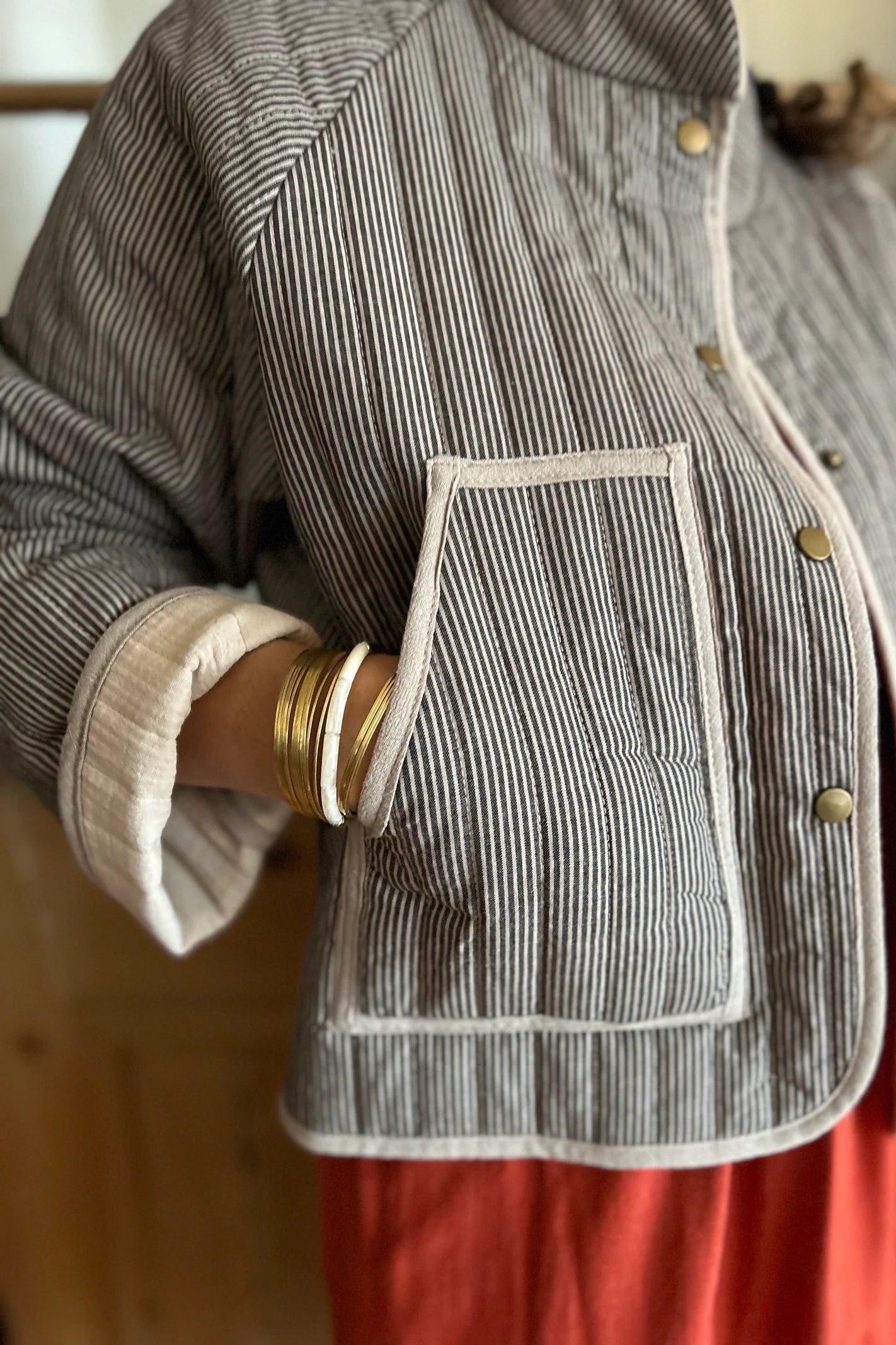 Close up of a woman wearing the Artemesia Jacket by Kazak in Taupe Stripe, a quilted jacket with snaps up the front and large patch pockets. She is wearing it over  the Lavendula Dress Paprika and standing in front of a wooden wall. 