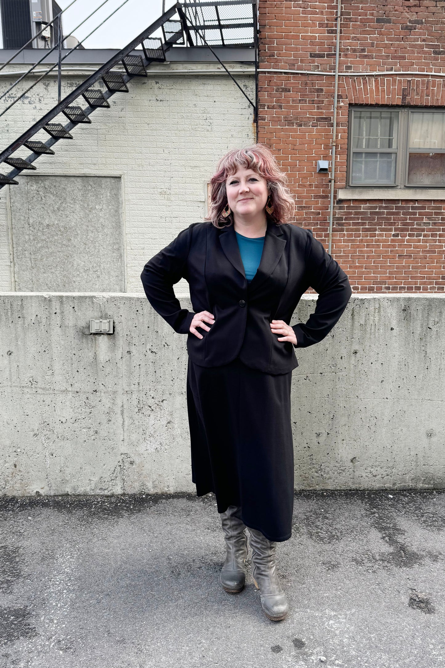 A woman wearing the Famke Jacket by Pure Essence in Black, a single breasted blazer with a single button closure. She is wearing it with a turquoise top and a black skirt and standing in front of a brick wall. 
