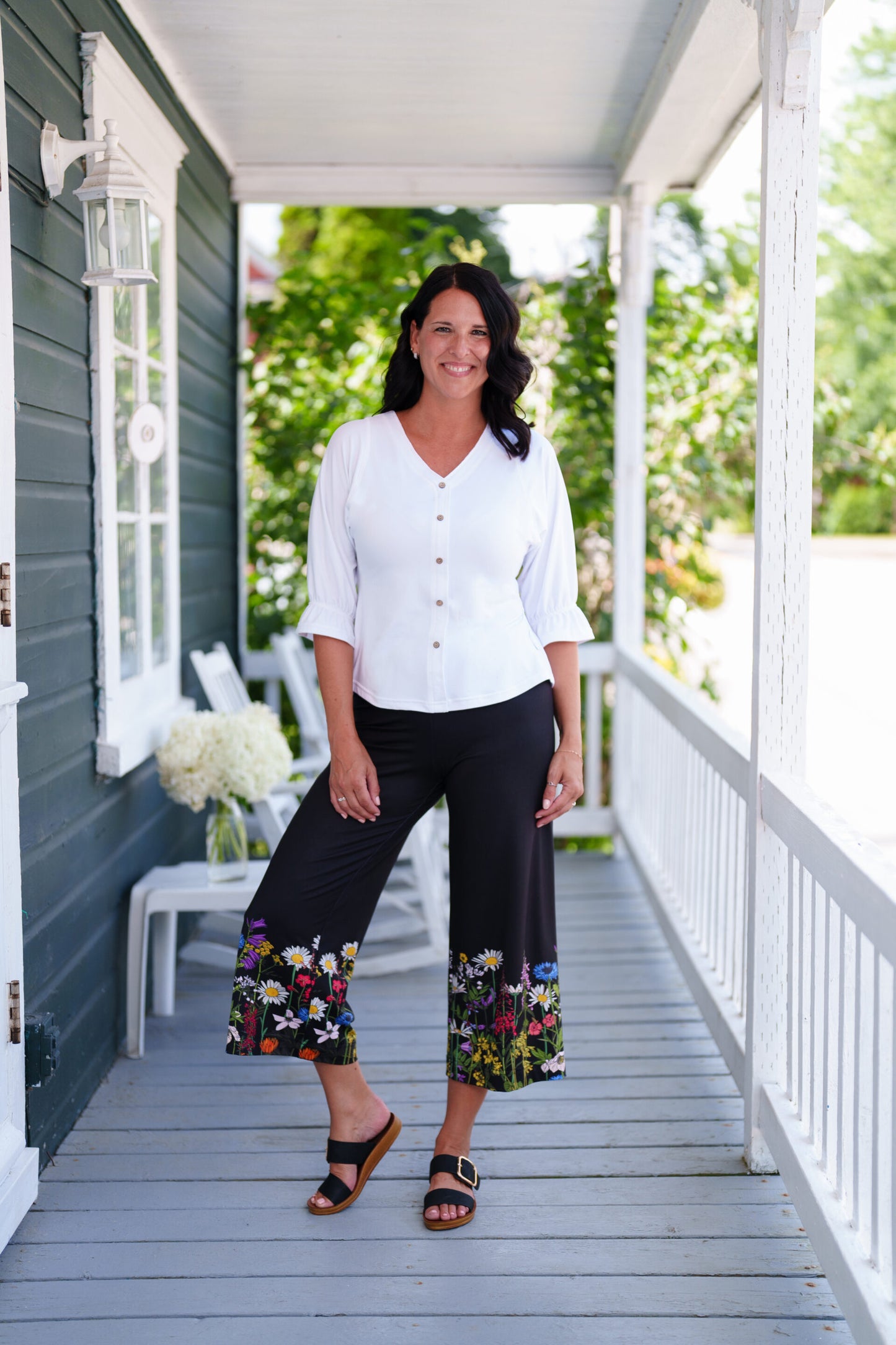 A woman wearing the Amaretto 7/8 Gaucho Pants from Rien ne se Perd in Black with a floral print hem. She is wearing them with a white top and standing on a porch. 