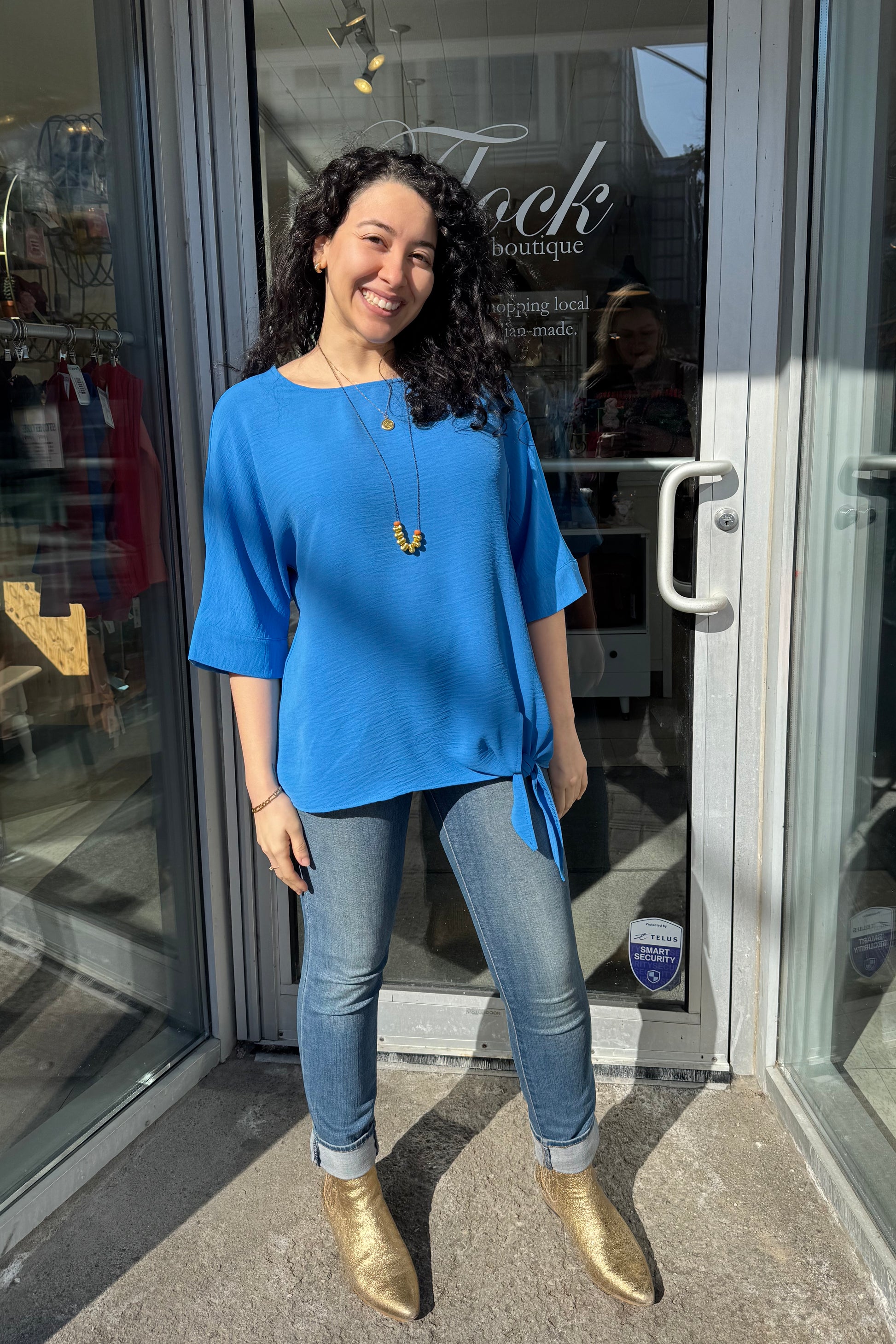 A woman wearing the Ginger Top from Pure Essence in Santorini blue, with a wide neck, blousy fit, 3/4 sleeves, and tie detail at the waist. She is wearing it with jeans and standing in the doorway at Flock. 