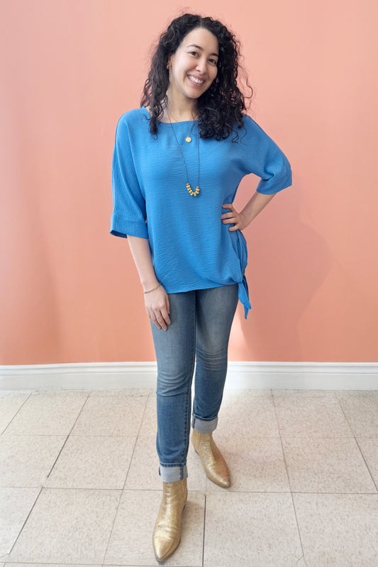 A woman wearing the Ginger Top from Pure Essence in Santorini blue, with a wide neck, blousy fit, 3/4 sleeves, and tie detail at the waist. She is wearing it with jeans  against a coral wall.