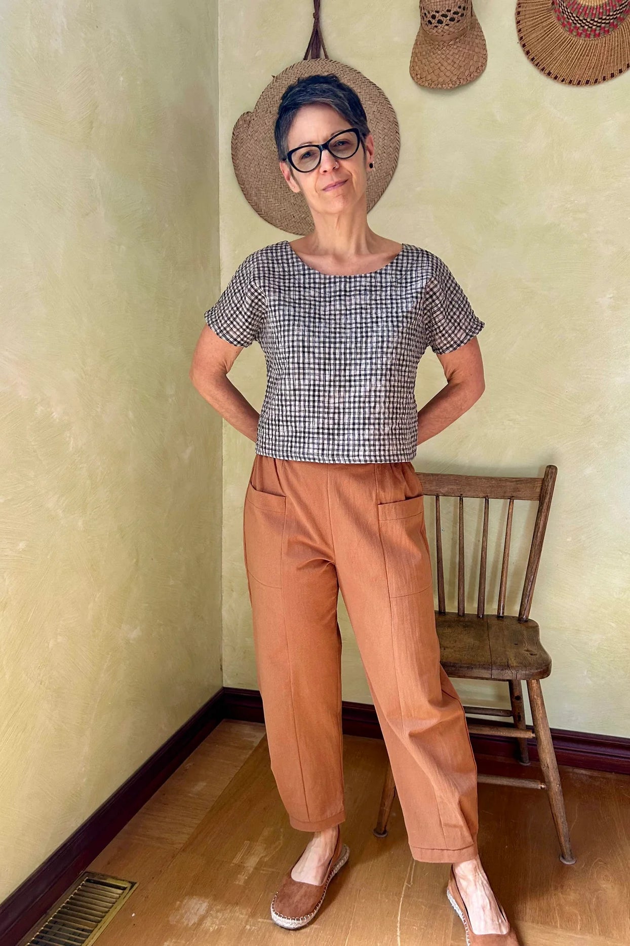 A woman wearing the Brendola Top from Kazak in Black and Cream Vichy/Gingham, a slightly cropped short sleeved top with three brass snaps that can be worn at the back or the front. She is wearing it with the Oxalis Pants in Ginger and standing in a room with a wooden chair. 