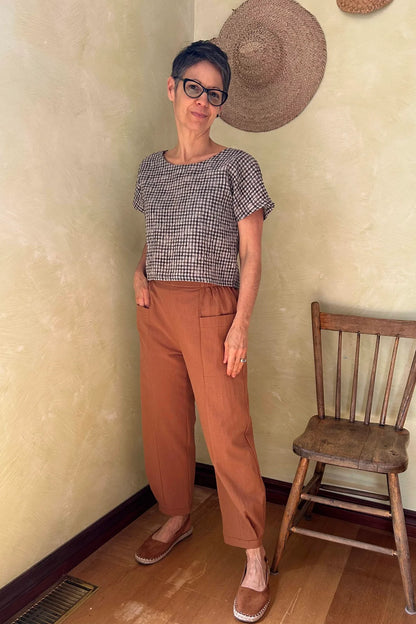 A woman wearing the Brendola Top from Kazak in Black and Cream Vichy/Gingham, a slightly cropped short sleeved top with three brass snaps that can be worn at the back or the front. She is wearing it with the Oxalis Pants in Ginger and standing in a room with a wooden chair. 
