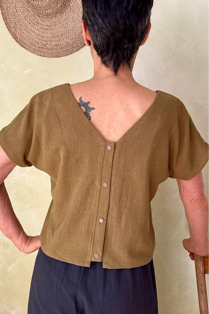 A woman wearing the Brendola Top from Kazak in Mousse, a slightly cropped short sleeved top with three brass snaps that can be worn at the back or the front. She is wearing it with the navy pants and standing in a room with hats on the wall. 
