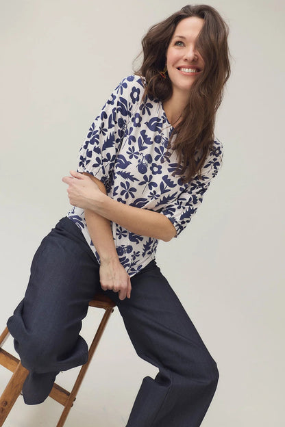 A woman wearing the Cadieux Top from message Factory in a floral Navy Print, with 3/4 sleeves with gathered cuffs, a wide neck and slim fit.  She is wearing it with dark pants and sitting on a stool against a plain background. 