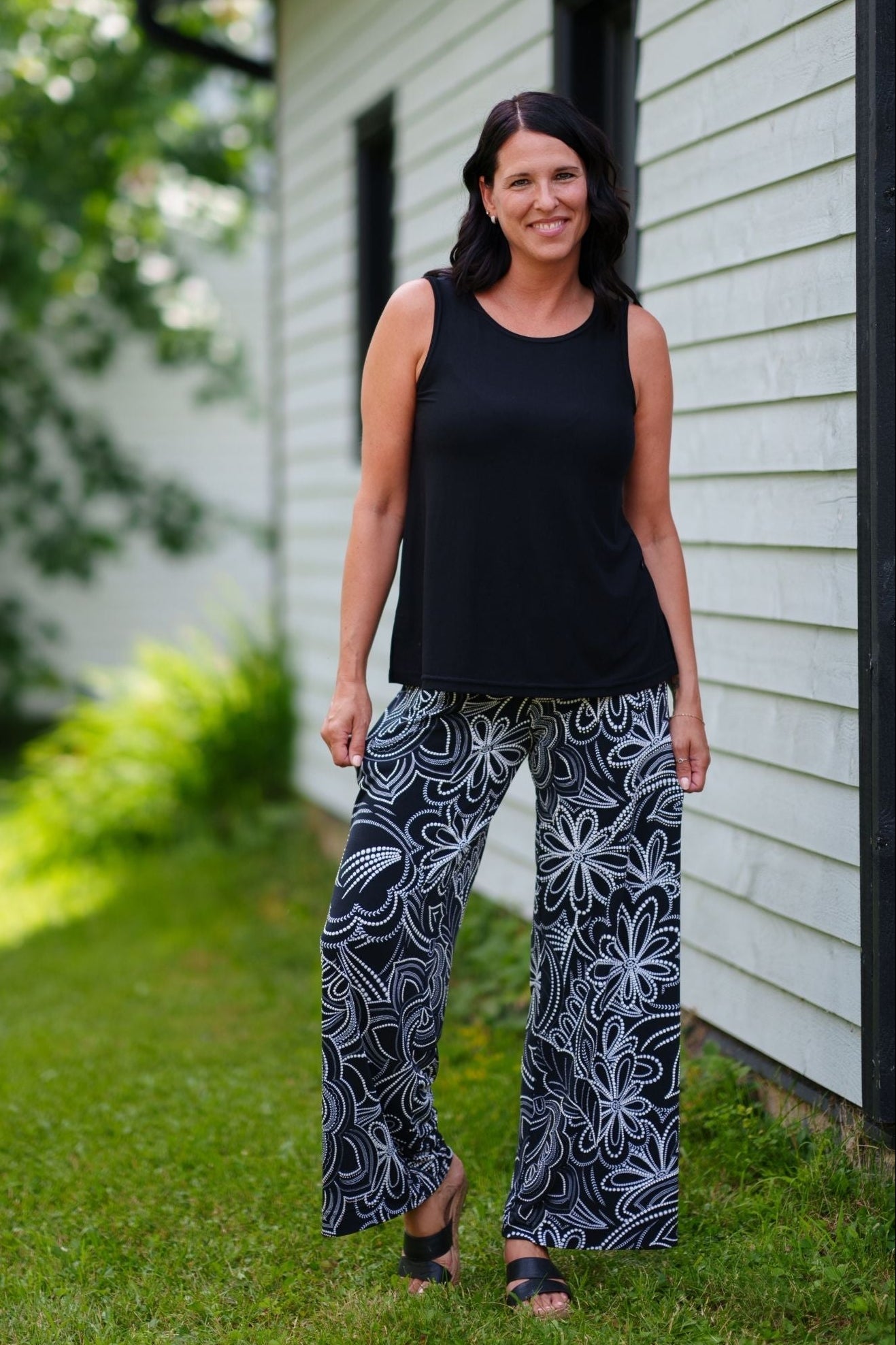 A woman wearing the Coco Palazzo Pants by Rien ne se Perd in Black and White floral, with a wide elastic waistband and wide legs. She is wearing them with a black top and standing outside by a wooden building. 