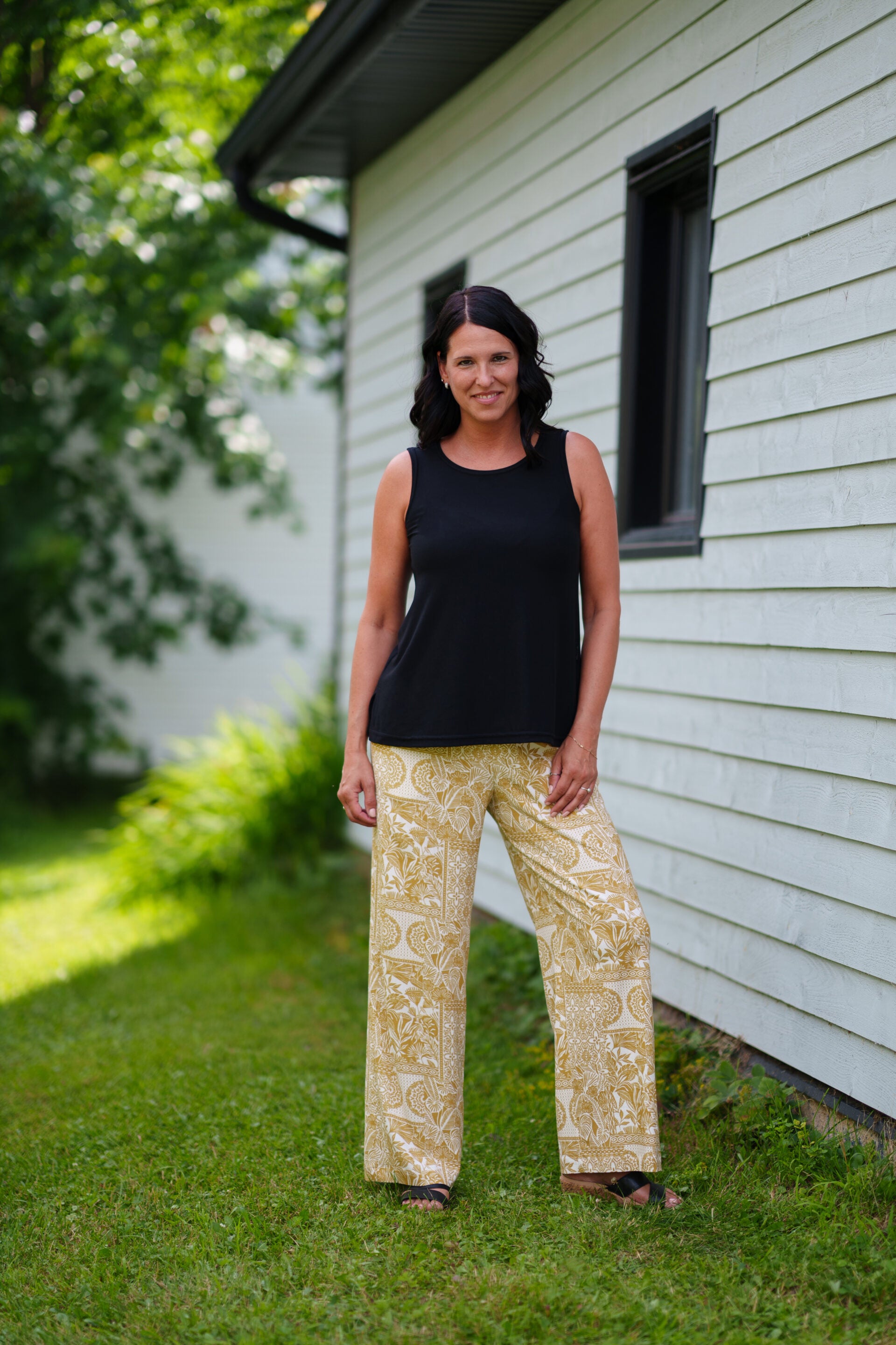 A woman wearing the Mango Pants from Rien ne se Perd in Ocre/Ivory tropical print, palazzo pants with an elastic waist and pockets. She is wearing them with a black top and standing on a porch. 