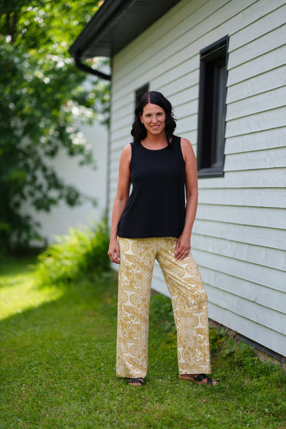 A woman wearing the Mango Pants from Rien ne se Perd in Ocre/Ivory tropical print, palazzo pants with an elastic waist and pockets. She is wearing them with a black top and standing on a porch. 