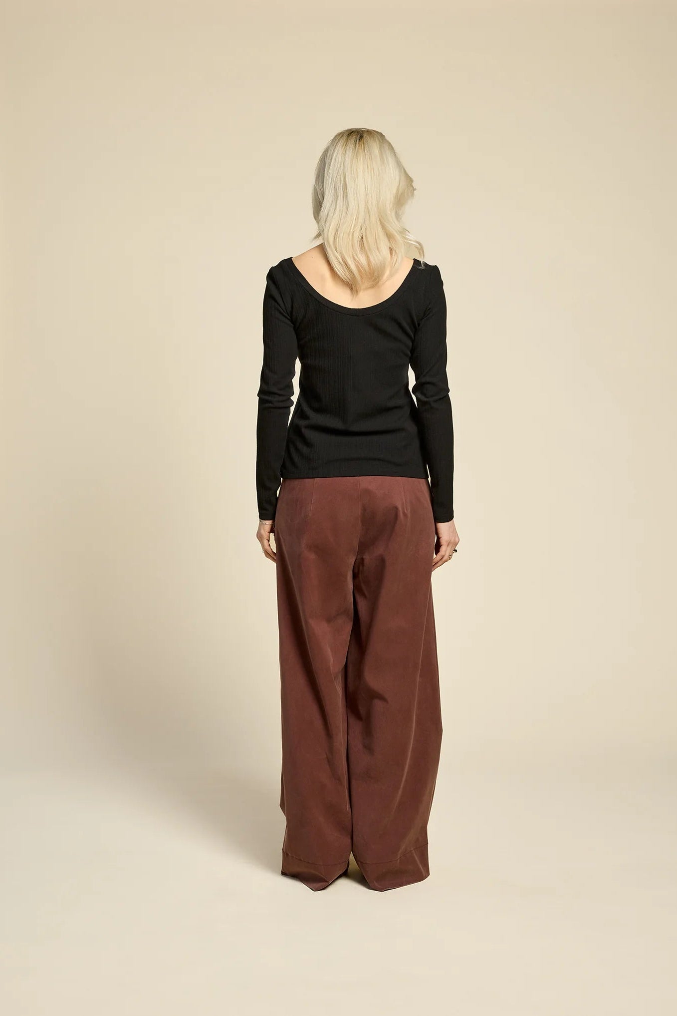 Back view of a woman wearing  the Chain pants from Cokluch in Cappuccino, with a slightly low waist, pointed belt loops, front pleats, and wide legs. She is wearing them with a black top  and standing against a grey background. 