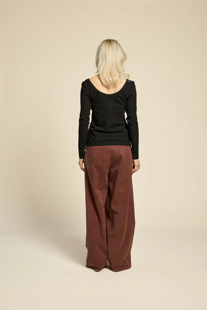 Back view of a woman wearing  the Chain pants from Cokluch in Cappuccino, with a slightly low waist, pointed belt loops, front pleats, and wide legs. She is wearing them with a black top  and standing against a grey background. 