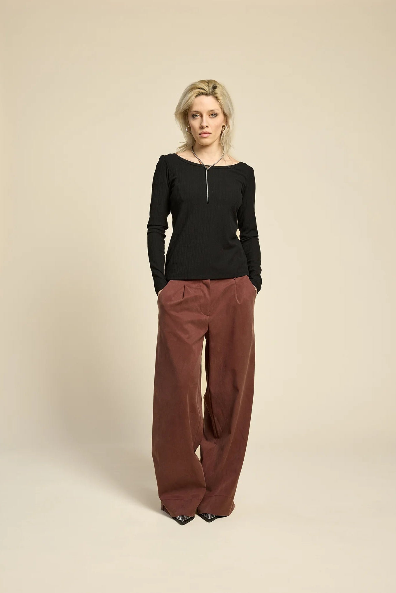 A woman wearing  the Chain pants from Cokluch in Cappuccino, with a slightly low waist, pointed belt loops, front pleats, and wide legs. She is wearing them with a black top  and standing against a grey background. 