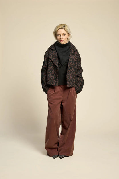 A woman wearing  the Chain pants from Cokluch in Cappuccino, with a slightly low waist, pointed belt loops, front pleats, and wide legs. She is wearing them with a leopard print jacket and standing against a grey background. 