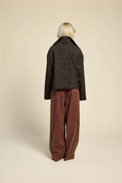 Back view of a woman wearing  the Chain pants from Cokluch in Cappuccino, with a slightly low waist, pointed belt loops, front pleats, and wide legs. She is wearing them with a leopard print jacket and standing against a grey background. 