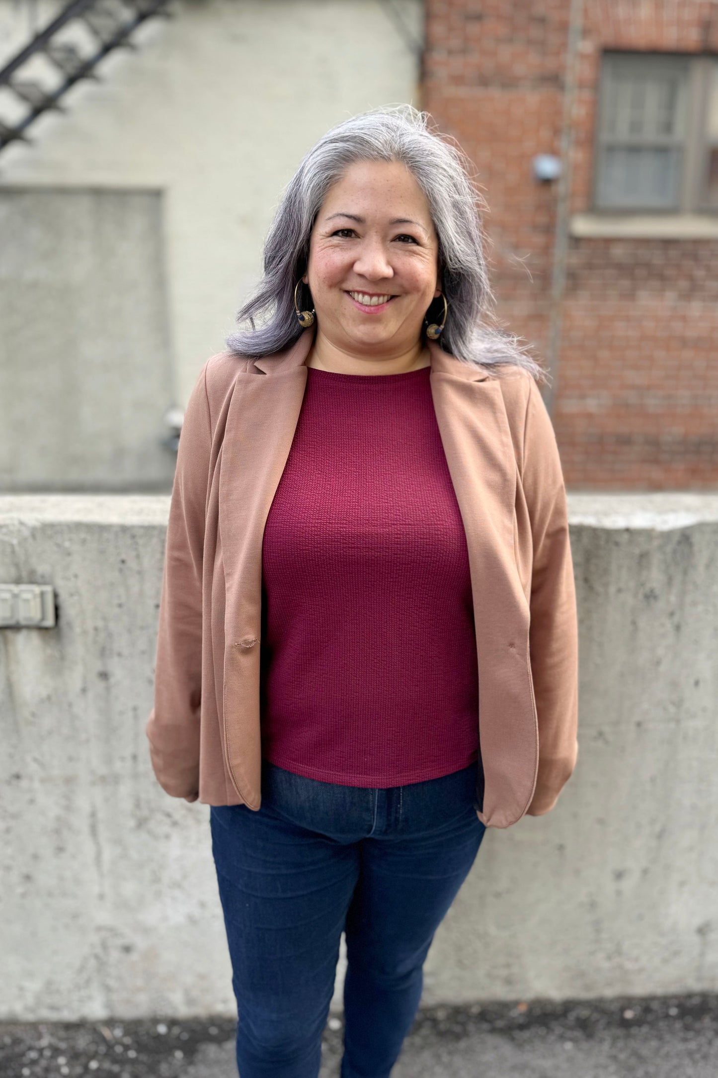A woman wearing the Famke Jacket by Pure Essence in Camel, a single breasted blazer with a single button closure. She is wearing it with jeans and a burgundy top and standing in front of a brick wall. 