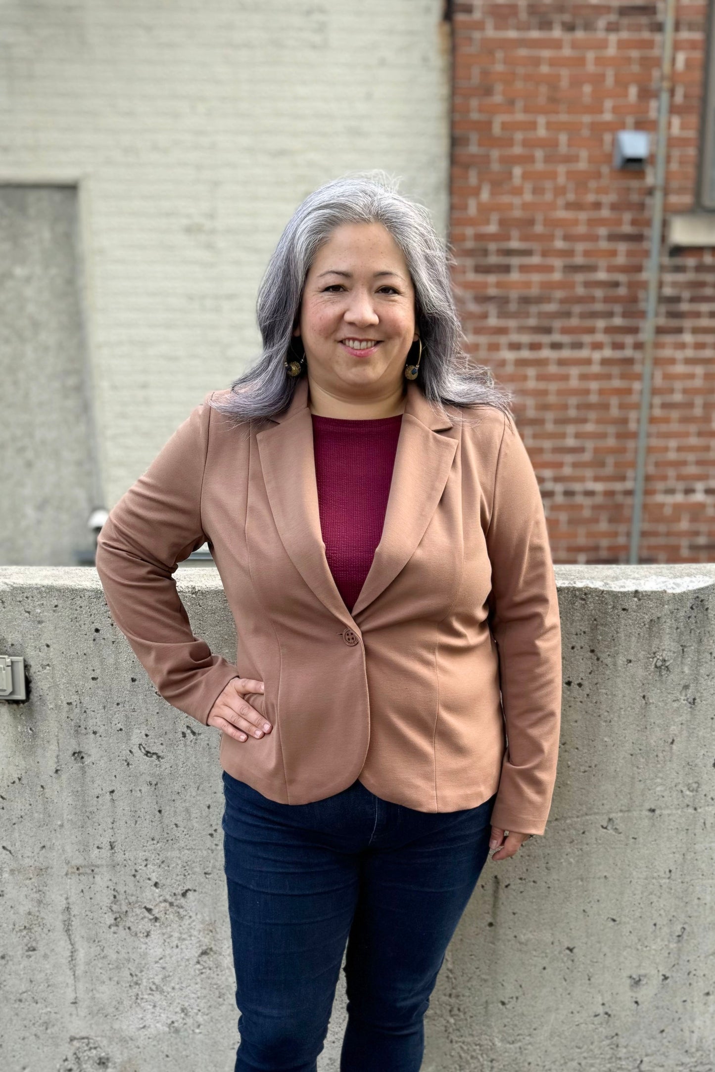 A woman wearing the Famke Jacket by Pure Essence in Camel, a single breasted blazer with a single button closure. She is wearing it with jeans and a burgundy top and standing in front of a brick wall. 