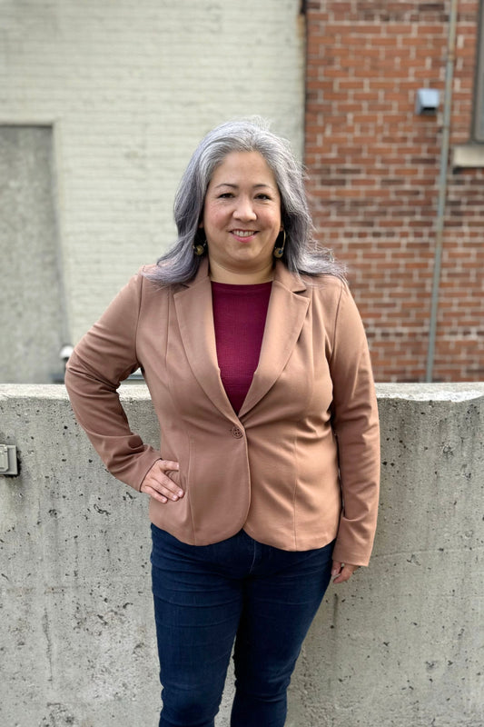A woman wearing the Famke Jacket by Pure Essence in Camel, a single breasted blazer with a single button closure. She is wearing it with jeans and a burgundy top and standing in front of a brick wall. 
