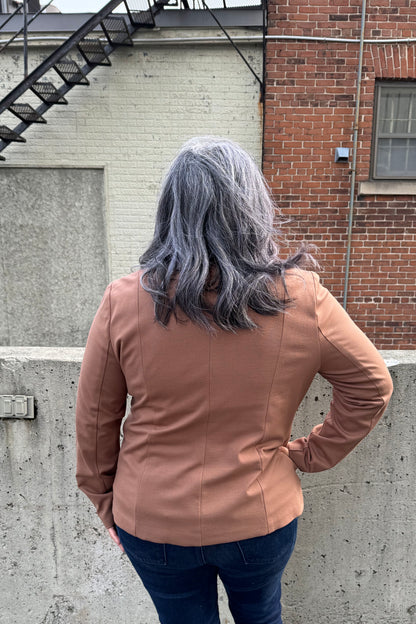 Back view of a woman wearing the Famke Jacket by Pure Essence in Camel, a single breasted blazer with a single button closure. She is wearing it with jeans and a burgundy top and standing in front of a brick wall. 