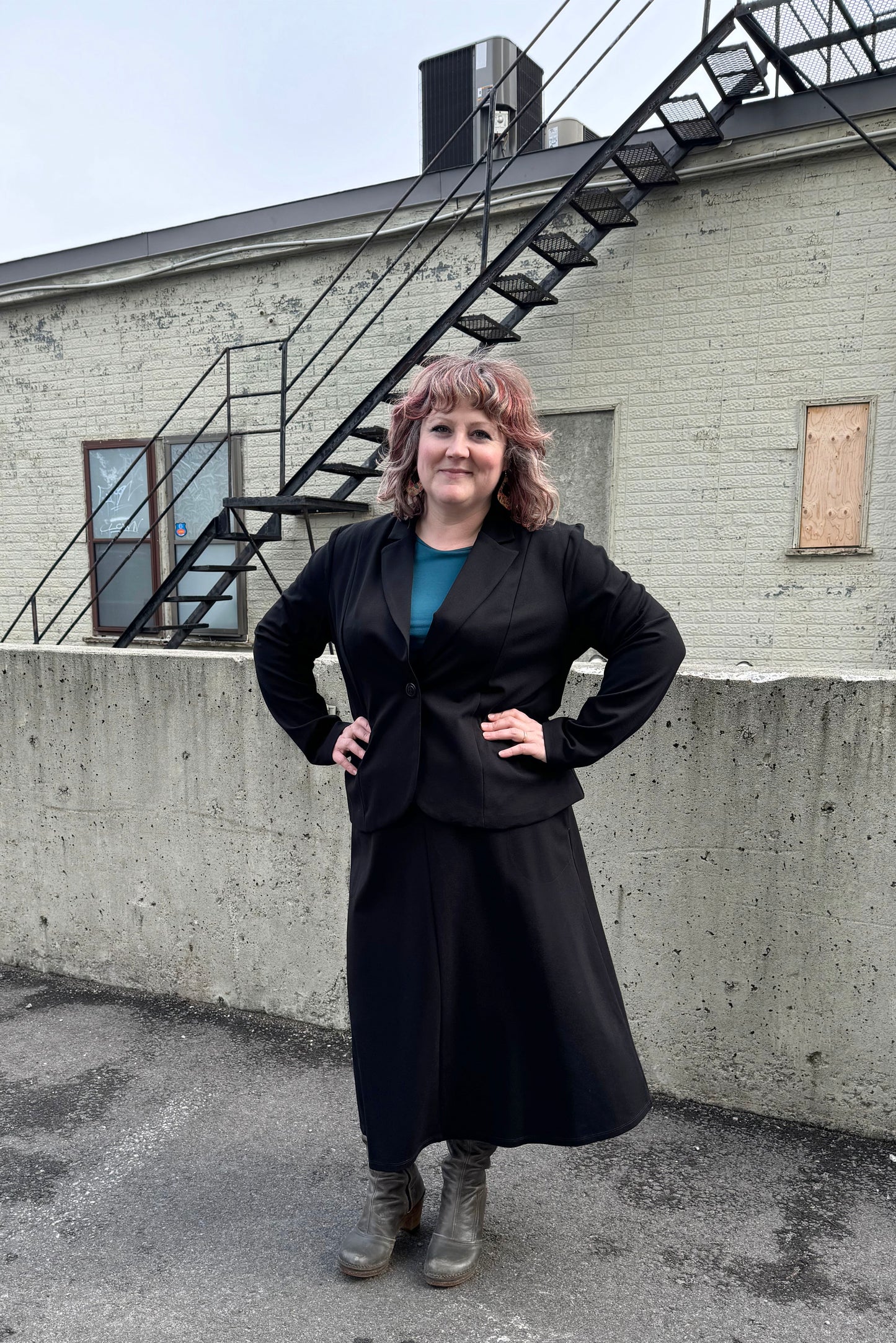 A woman wearing the Famke Jacket by Pure Essence in Black, a single breasted blazer with a single button closure. She is wearing it with a turquoise top and a black skirt and standing in front of a brick wall. 