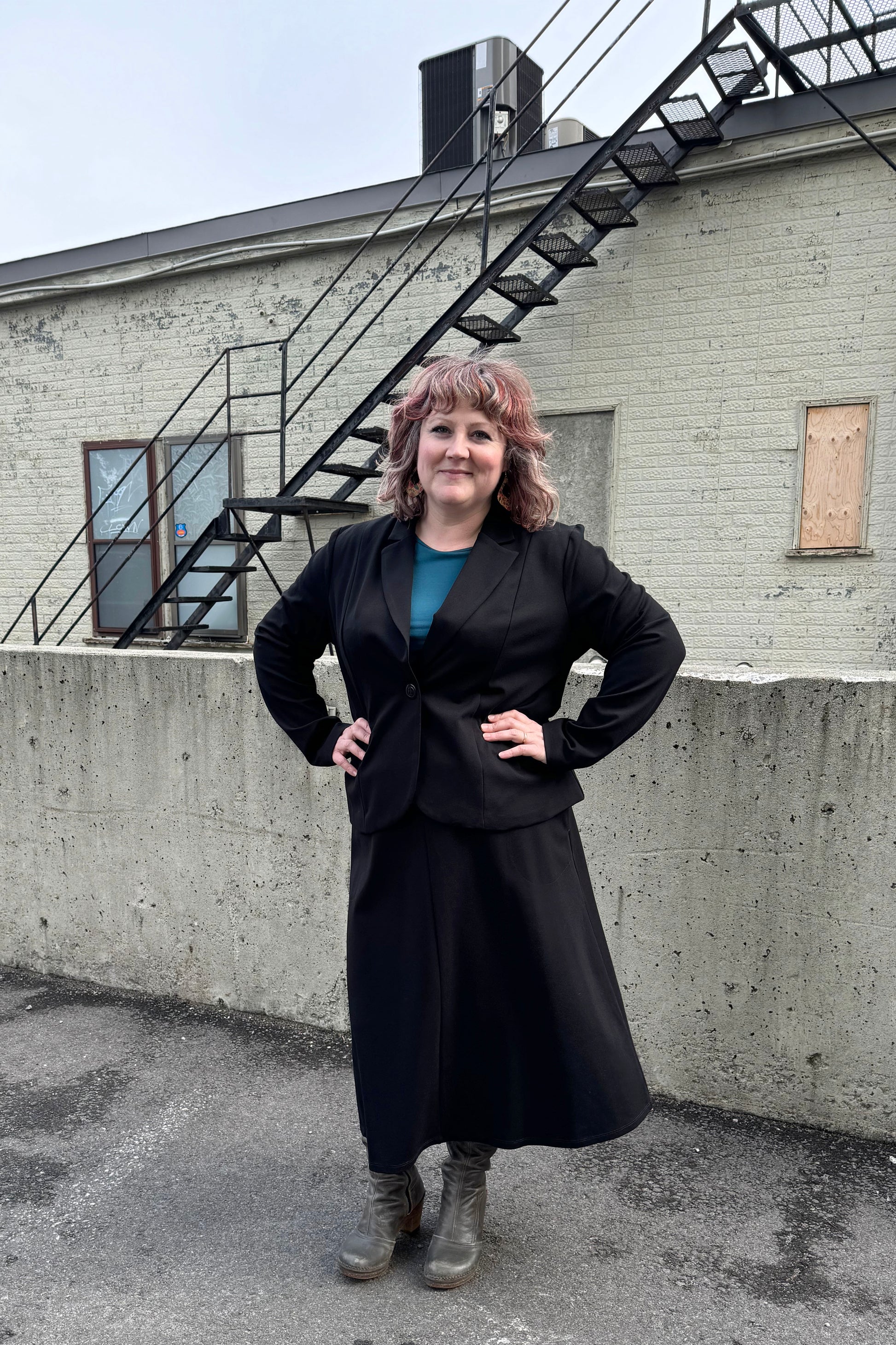 A woman wearing the Famke Jacket by Pure Essence in Black, a single breasted blazer with a single button closure. She is wearing it with a turquoise top and a black skirt and standing in front of a brick wall. 