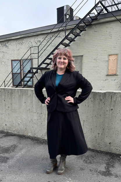 A woman wearing the Famke Jacket by Pure Essence in Black, a single breasted blazer with a single button closure. She is wearing it with a turquoise top and a black skirt and standing in front of a brick wall. 