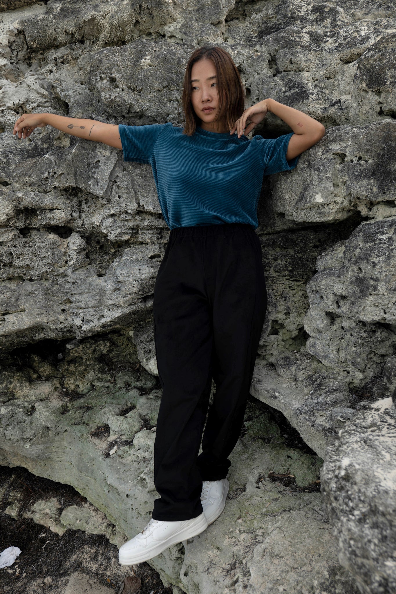 A woman wearing the Funchal Pants by MAS in Pepper, straight legged corduroy pants with an elastic waist. she is wearing them with a blue velvet top and standing in front of a rock wall. 