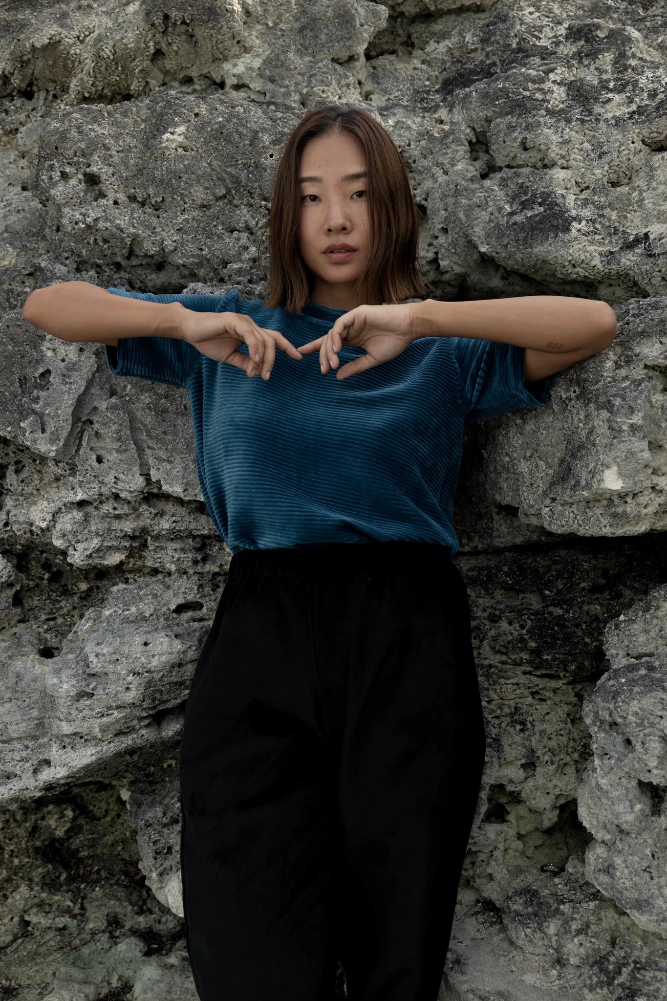 A woman wearing the Funchal Pants by MAS in Pepper, straight legged corduroy pants with an elastic waist. she is wearing them with a blue velvet top and standing in front of a rock wall. 