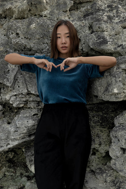 A woman wearing the Funchal Pants by MAS in Pepper, straight legged corduroy pants with an elastic waist. she is wearing them with a blue velvet top and standing in front of a rock wall. 