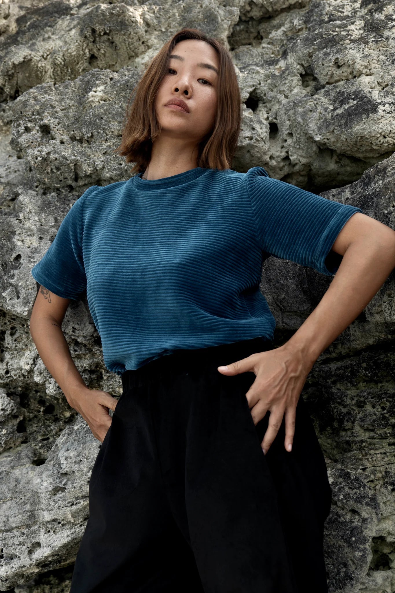 A woman wearing the Funchal Pants by MAS in Pepper, straight legged corduroy pants with an elastic waist. she is wearing them with a blue velvet top and standing in front of a rock wall. 