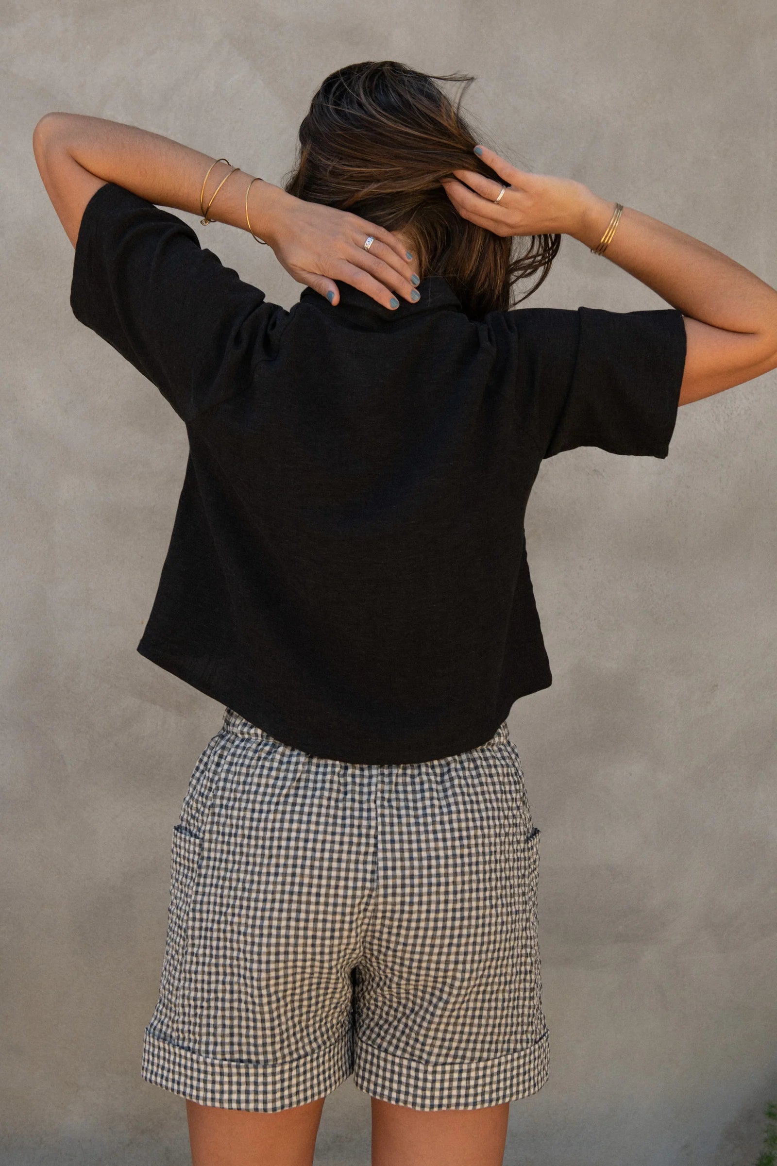 Back view of a woman wearing the Hosta Screen-printed Blouse by Kazak in Black with Dandelions, with hand-screen-printed flowers, mid-length raglan sleeves, a classic collar, and a button front. She is wearing it with gingham shorts and standing outside by a grey wall. 