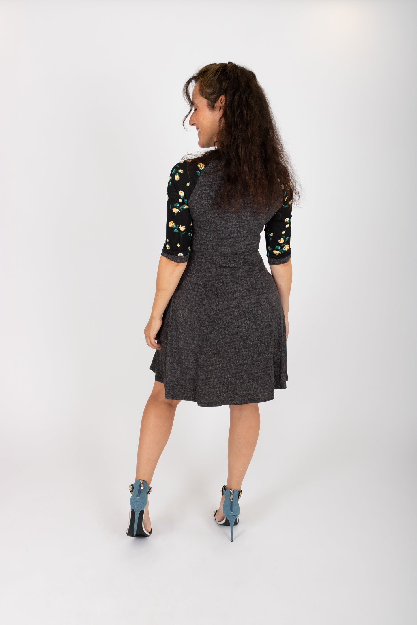 Back view of a woman wearing the Raphaelle Dress from Infime in Black, an A-line knee-length dress with 3/4 raglan sleeves that feature a floral print and pockets with a floral detail. She is standing in front of a white background