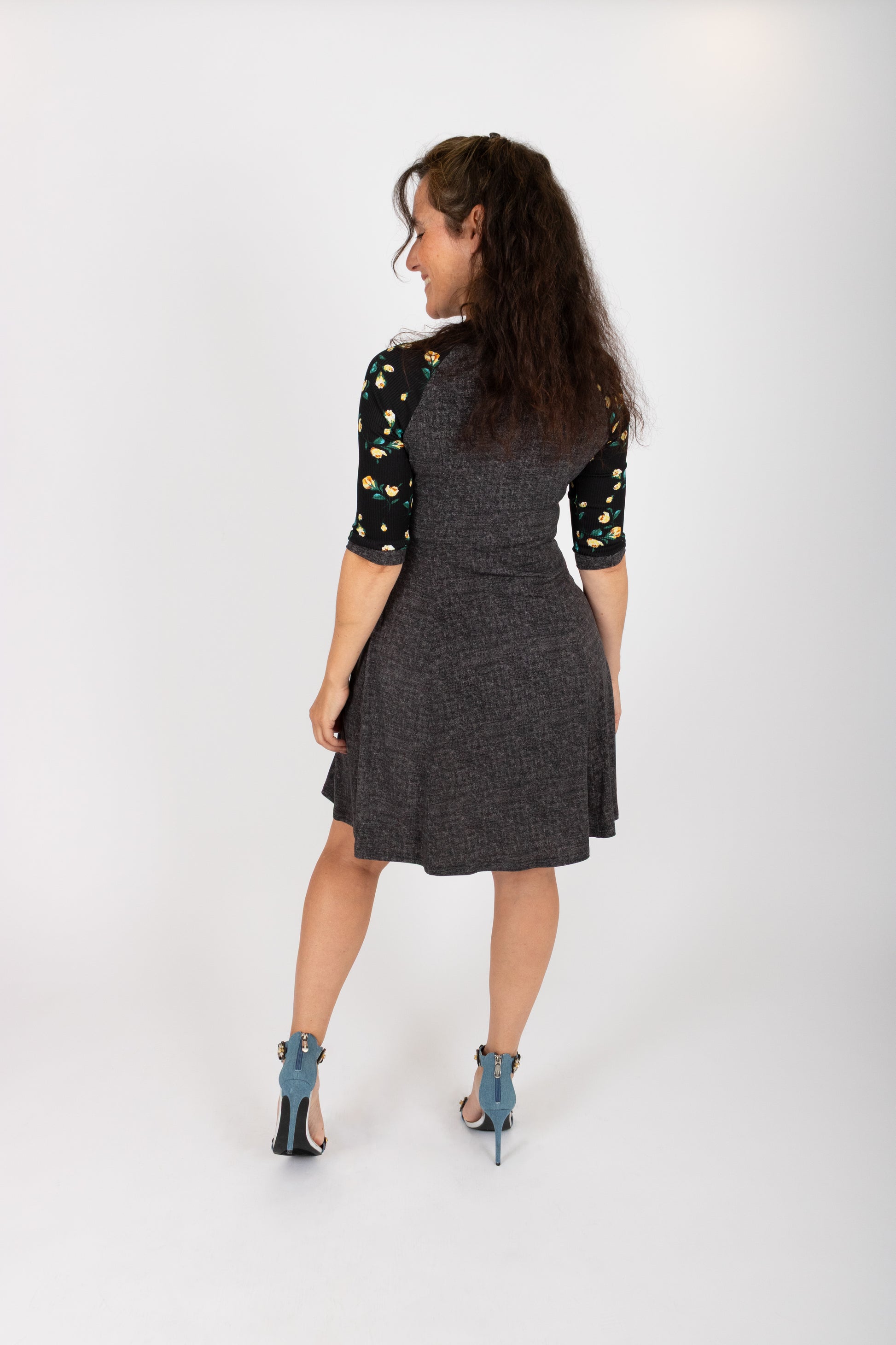 Back view of a woman wearing the Raphaelle Dress from Infime in Black, an A-line knee-length dress with 3/4 raglan sleeves that feature a floral print and pockets with a floral detail. She is standing in front of a white background