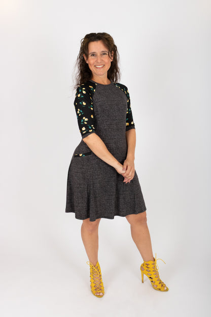 A woman wearing the Raphaelle Dress from Infime in Black, an A-line knee-length dress with 3/4 raglan sleeves that feature a floral print and pockets with a floral detail. She is standing in front of a white background