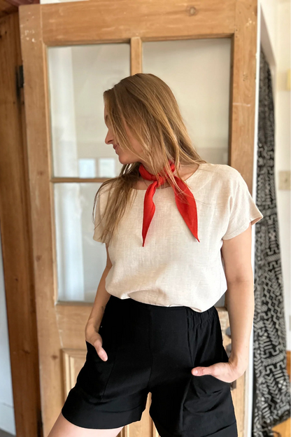 A woman wearing the Iris Shorts by Kazak in Black, with a smooth front waist, elastic at the back, large pockets, and mid-thigh length. She is wearing them with a cream top and red scarf and standing in a room with a door. 