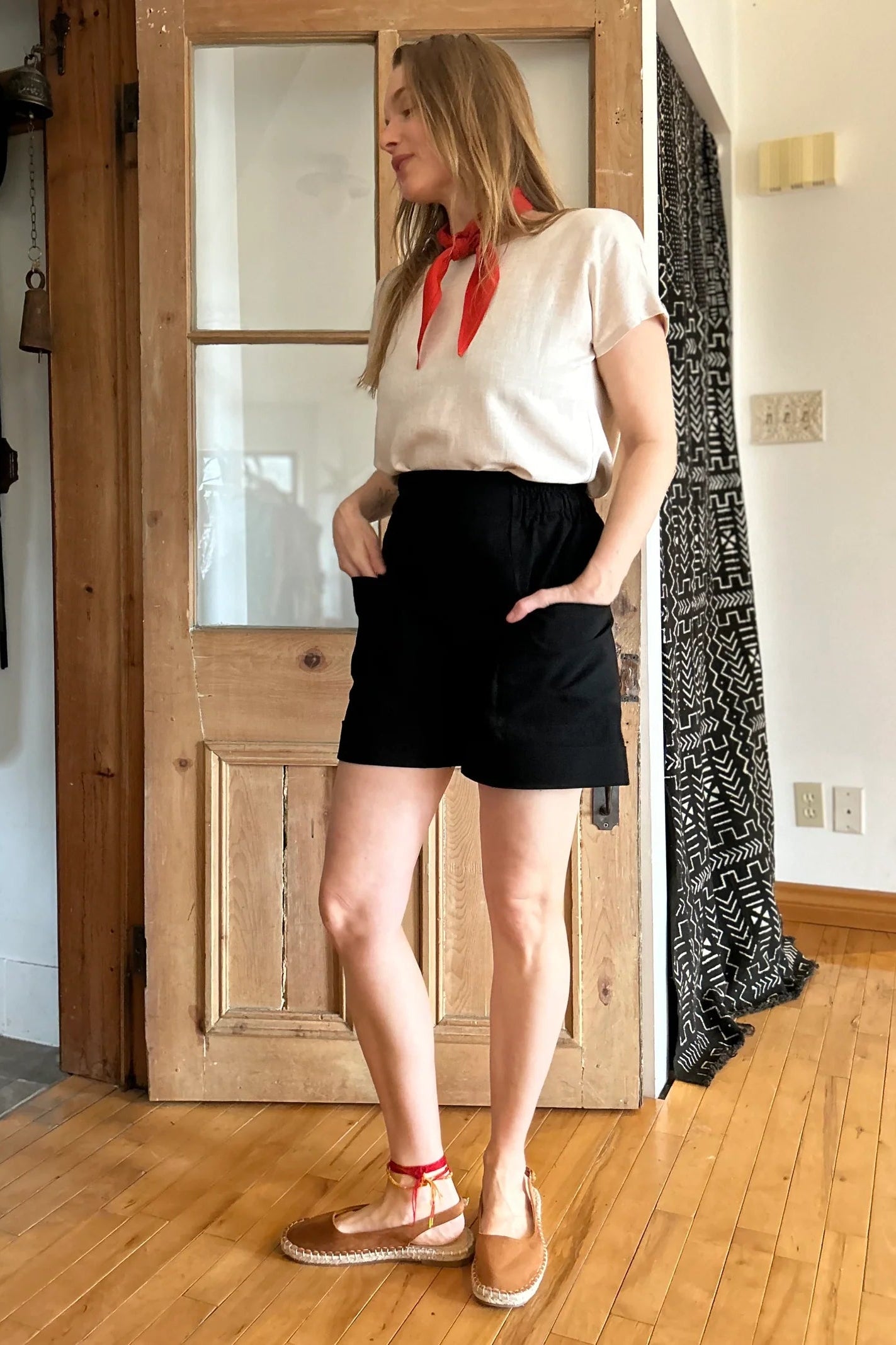 A woman wearing the Iris Shorts by Kazak in Black, with a smooth front waist, elastic at the back, large pockets, and mid-thigh length. She is wearing them with a cream top and red scarf and standing in a room with a door. 