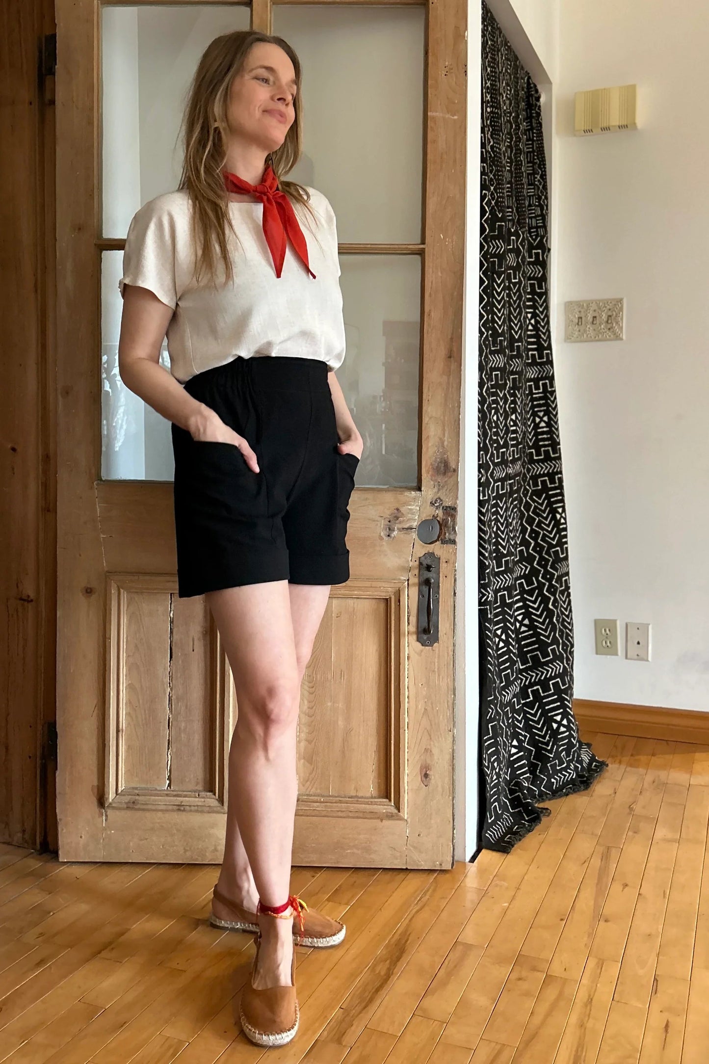 A woman wearing the Iris Shorts by Kazak in Black, with a smooth front waist, elastic at the back, large pockets, and mid-thigh length. She is wearing them with a cream top and red scarf and standing in a room with a door. 