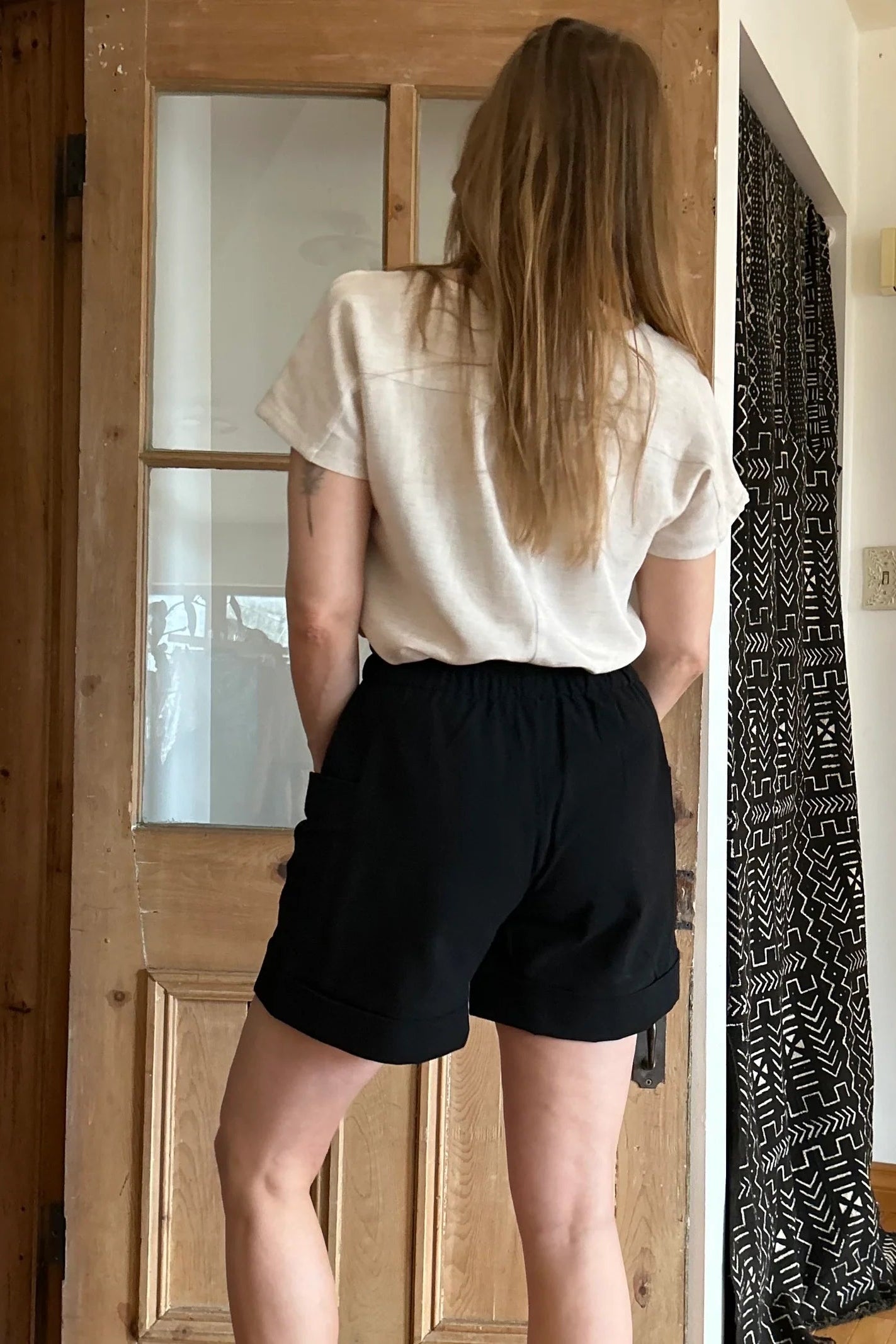 Back view of a woman wearing the Iris Shorts by Kazak in Black, with a smooth front waist, elastic at the back, large pockets, and mid-thigh length. She is wearing them with a cream top and red scarf and standing in a room with a door. 