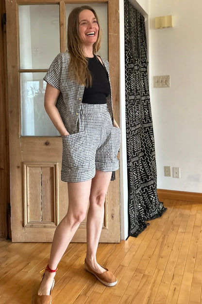 A woman wearing the Iris Shorts by Kazak in Black and Cream Vichy/Gingham, with a smooth front waist, elastic at the back, large pockets, and mid-thigh length. She is wearing them with a black tank and matching open top and standing in a room with a door. 