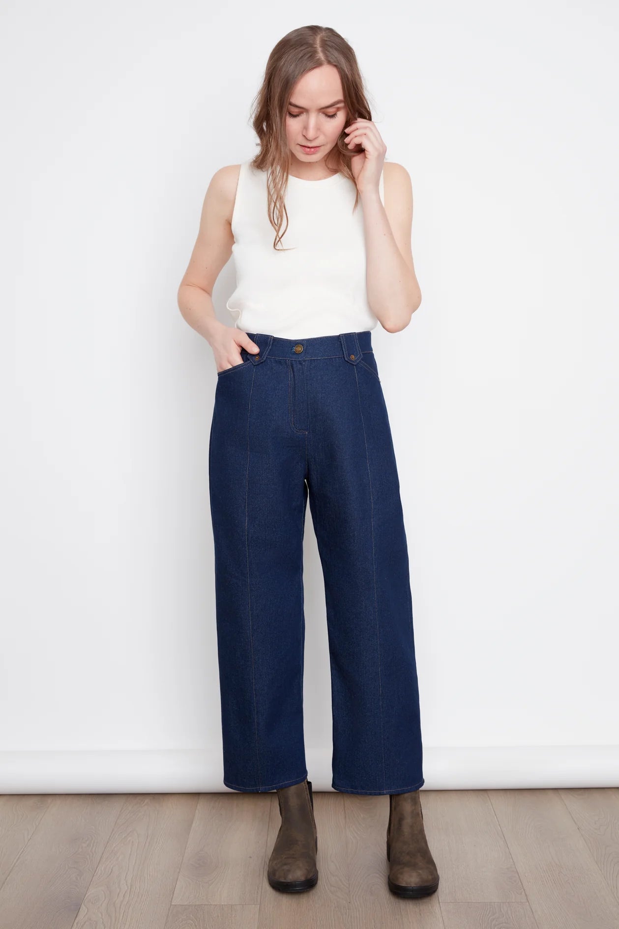 A woman wearing the Alice Straight Leg Jeans by Julei in Dark Denim, with an elastic back waistband, seams at the front and back of the legs, and pointed belt loops. She is wearing them with a white tank and standing in front of a white background. 