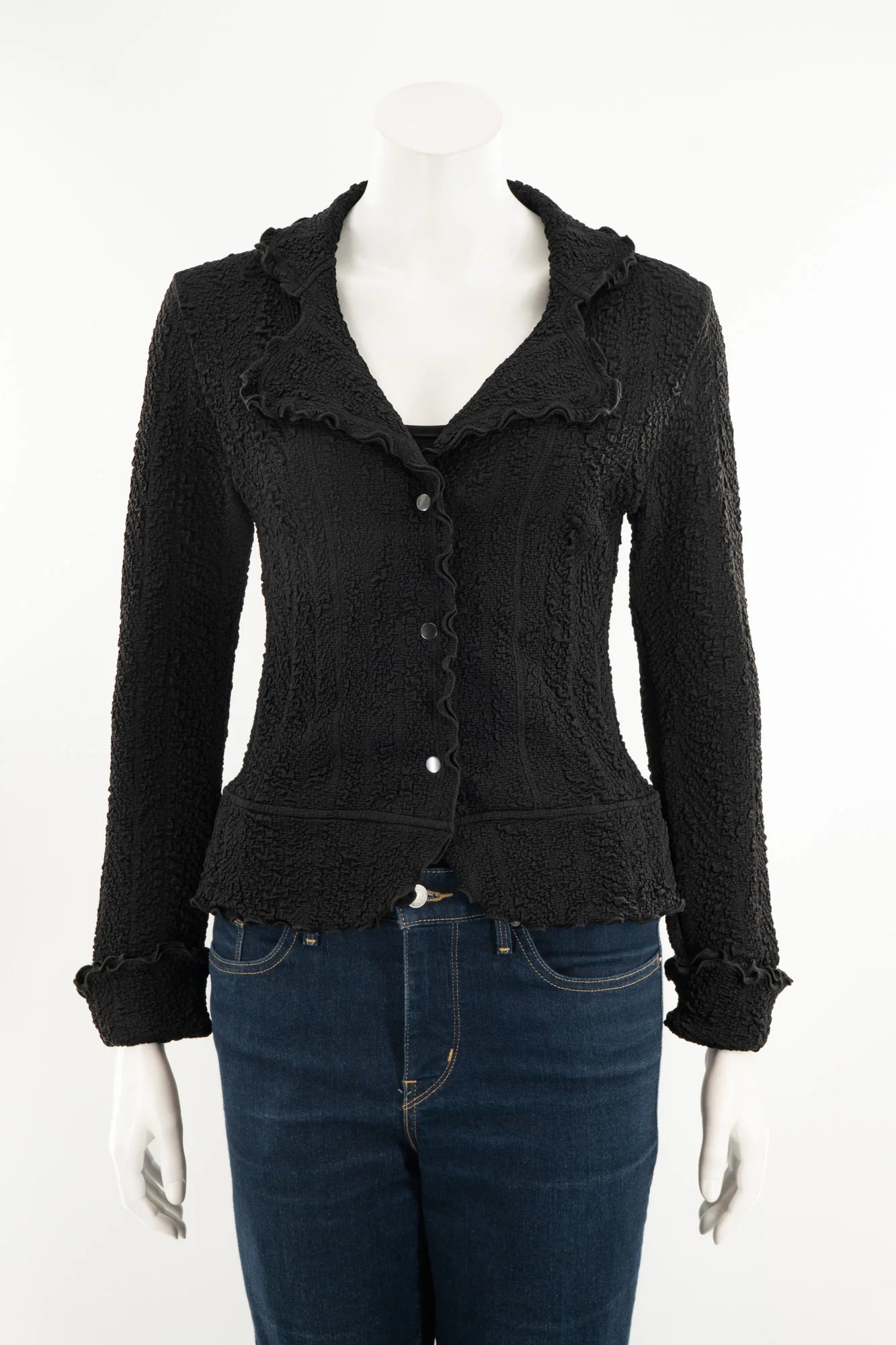 The Essential Jacket - Jasper by Karkass is shown on a mannequin in front of a white background. It's a black textured jacket with a ruffled collar, button closure, and frill details at the cuffs and hem. 