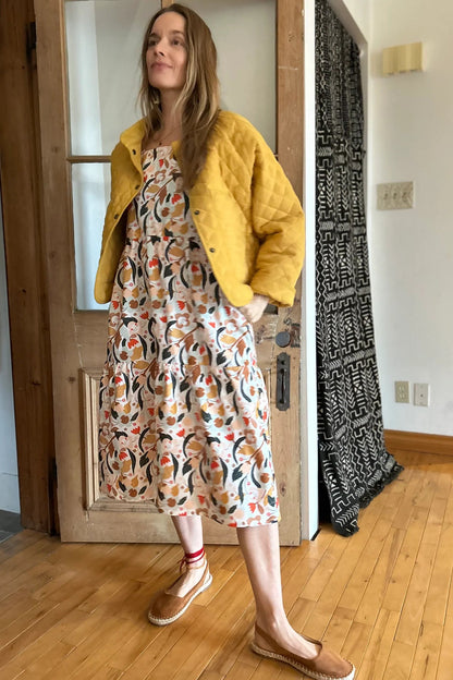 Woman wearing the Lavendula Dress by Kazak in Floral with cream, a short sleeved square neck midi dress with three ruffles. She is wearing it with a yellow jacket and standing in a room with wooden floors and a door.
