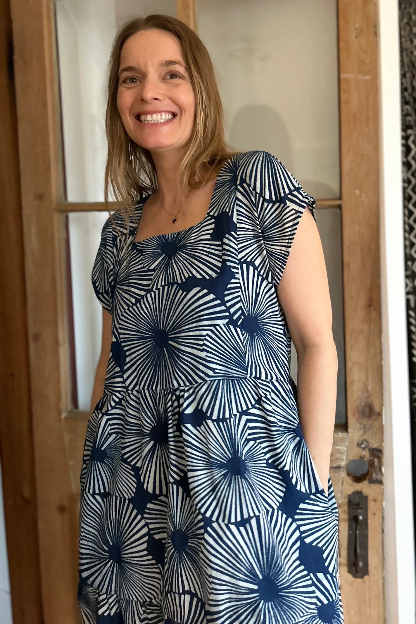 Woman wearing the Lavendula Dress by Kazak in Floral Indigo, a short sleeved square neck midi dress with three ruffles. She is standing in a room with wooden floors and a door.