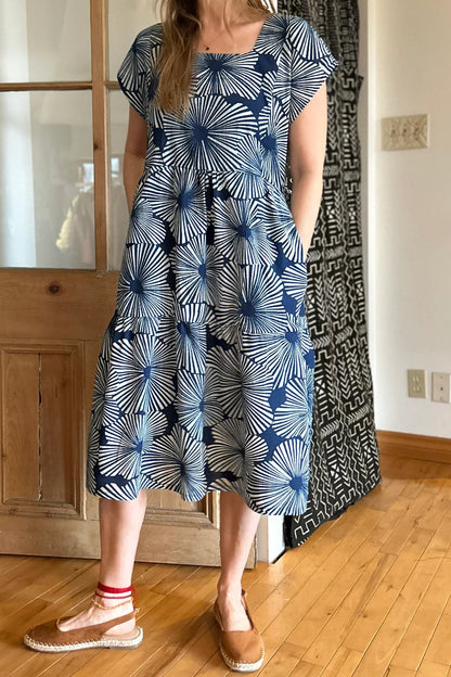 Woman wearing the Lavendula Dress by Kazak in Floral Indigo, a short sleeved square neck midi dress with three ruffles. She is standing in a room with wooden floors and a door.