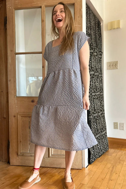 A woman wearing the Lavendula Dress by Kazak in Sky Vichy/Gingham, a short sleeved square neck midi dress with three ruffles. She is standing in a room with wooden floors and a door.