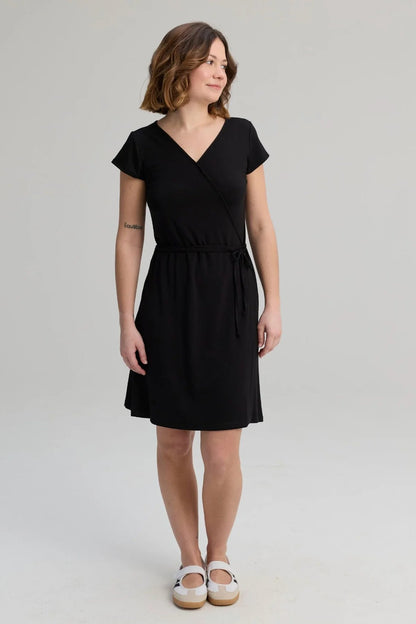 A woman wearing the Libellule Dress by Message Factor in Black, a short sleeved wrap dress that falls to the knee. She is standing against a plain background. 